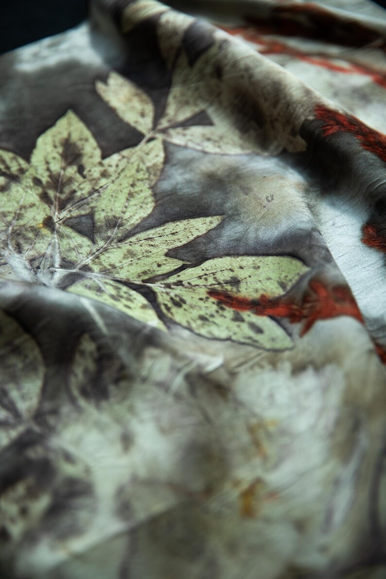 May include: A lightweight fabric featuring a botanical print with large, detailed green leaves and dark brown, mottled background tones. The textile displays organic, earthy patterns with subtle accents of deep red throughout the surface.