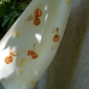 May include: A white fabric scarf featuring a botanical print with scattered, rust-colored circular leaf or seed pod patterns. The material is lightweight and semi-translucent, showing natural folds and soft draping as it hangs vertically.