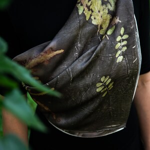May include: A dark brown scarf featuring a botanical print of light green fern-like leaves and foliage patterns. The fabric has a textured appearance with organic shapes and natural color variations across the surface of the draped material.