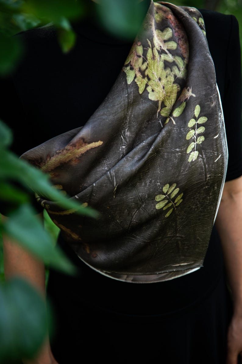 May include: A dark brown scarf featuring a botanical print of light green fern-like leaves and foliage patterns. The fabric has a textured appearance with organic shapes and natural color variations across the surface of the draped material.