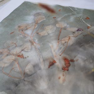 May include: A square textile piece featuring a botanical print with muted gray and green tones. The fabric displays a natural leaf and stem pattern in soft, faded shades of beige and brown, created through a direct plant-based dye process.