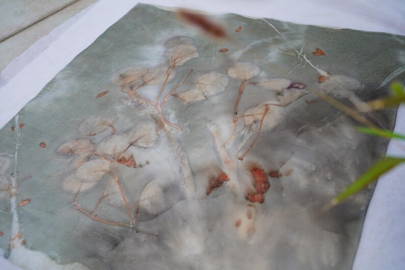 May include: A square textile piece featuring a botanical print with muted gray and green tones. The fabric displays a natural leaf and stem pattern in soft, faded shades of beige and brown, created through a direct plant-based dye process.