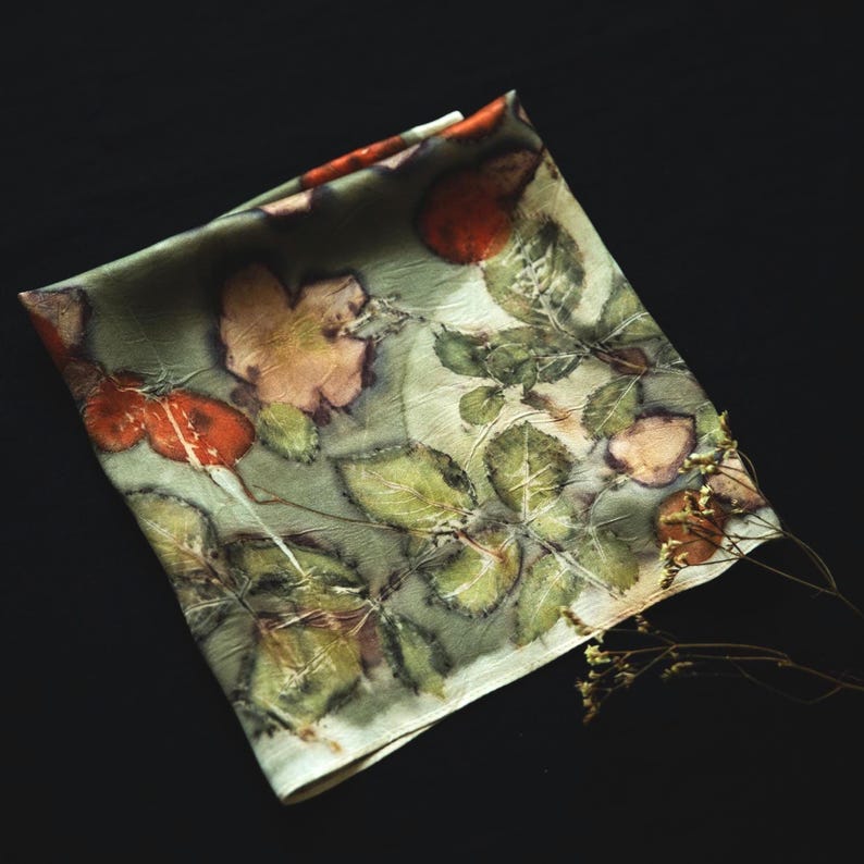 May include: A square silk scarf featuring a botanical print with muted green foliage, detailed leaf veins, and vibrant orange-red rose hips. The fabric has a soft, slightly wrinkled texture with a natural, organic pattern of plant life.