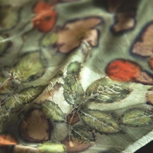 May include: A lightweight scarf featuring a botanical print with detailed green leaves and reddish-brown circular shapes on a pale sage green background. The fabric has a soft, slightly reflective sheen and displays natural folds and creases throughout.