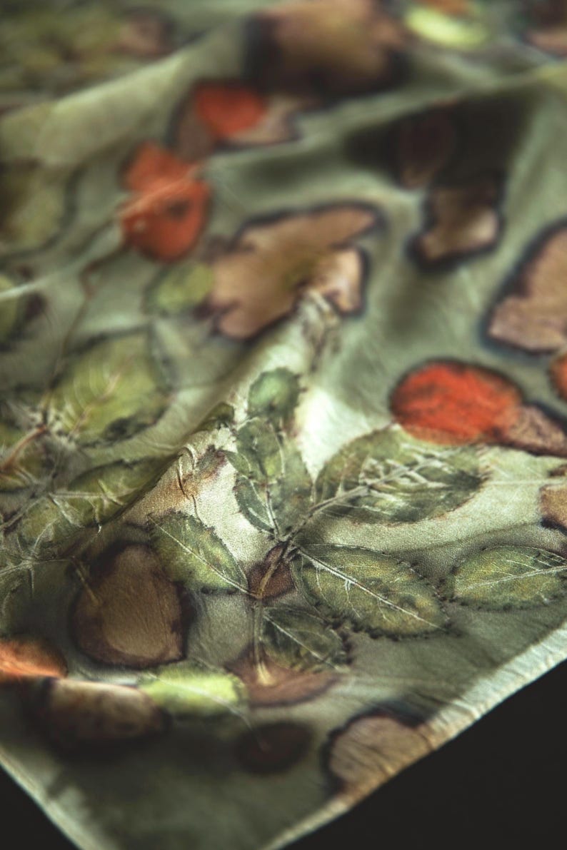 May include: A lightweight scarf featuring a botanical print with detailed green leaves and reddish-brown circular shapes on a pale sage green background. The fabric has a soft, slightly reflective sheen and displays natural folds and creases throughout.