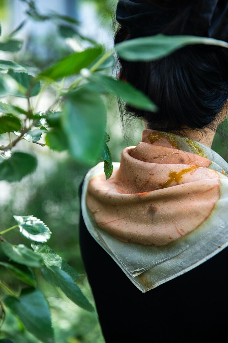 May include: A square scarf featuring a soft peach and cream color palette with organic, botanical-inspired leaf imprints in shades of rust and brown. The fabric has a delicate, semi-sheer texture and is folded around the neck in a layered style.