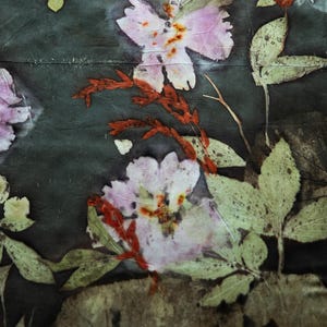 May include: A dark gray fabric featuring a botanical print with pressed light pink flowers, vibrant green leaves, and slender reddish-brown stems. The natural plant patterns are distributed across the surface, creating a detailed floral composition.