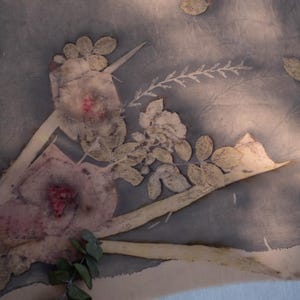 May include: A textile fabric featuring a botanical print with muted gray and blue tones. The design displays pressed flowers, leaves, and stems in shades of cream, pale pink, and soft green, creating a natural, layered floral pattern.