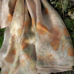 May include: A light beige scarf featuring a botanical print with various leaf shapes in shades of orange, brown, and muted green. The fabric has a smooth, soft texture with visible leaf vein patterns imprinted across the surface of the material.