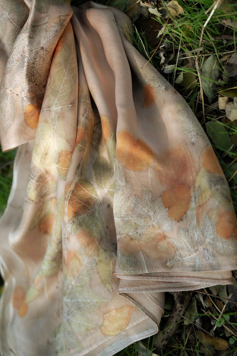 May include: A light beige scarf featuring a botanical print with various leaf shapes in shades of orange, brown, and muted green. The fabric has a smooth, soft texture with visible leaf vein patterns imprinted across the surface of the material.