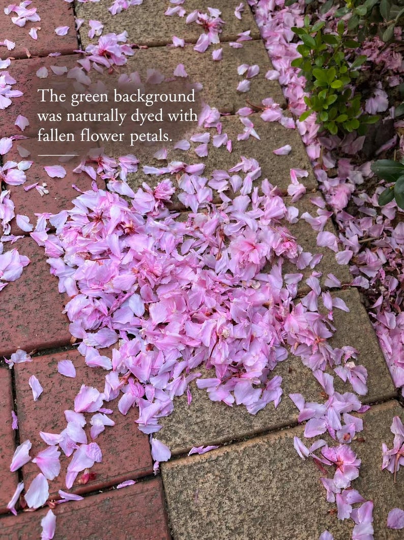 May include: A collection of light pink flower petals scattered across a paved surface of red and gray rectangular stones. The text overlay reads: The green background was naturally dyed with fallen flower petals. Green foliage is visible.