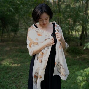 以下が含まれることがあります： 女性が、茶色とオレンジ色の花柄模様の、透け感のあるクリーム色のスカーフを着用しています。 スカーフは黒いドレスの上にドレープされており、柔らかく流れるようなシルエットを作り出しています。 背景はぼやけた森です。
