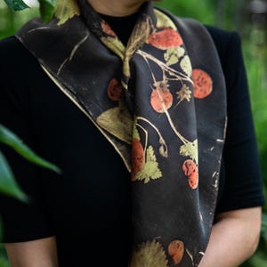 May include: A square scarf featuring a dark charcoal background with a botanical print of orange-red berries and pale green leaves. The fabric displays a natural, organic pattern of stems and foliage arranged across the surface of the accessory.