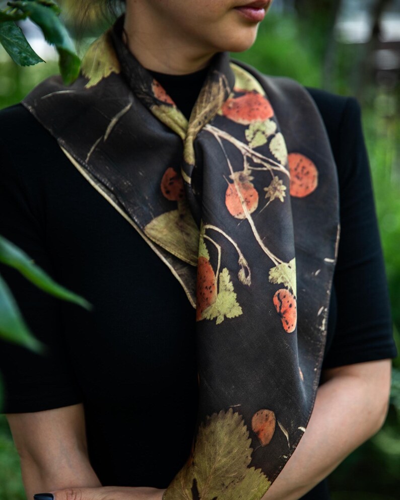 May include: A square scarf featuring a dark charcoal background with a botanical print of orange-red berries and pale green leaves. The fabric displays a natural, organic pattern of stems and foliage arranged across the surface of the accessory.