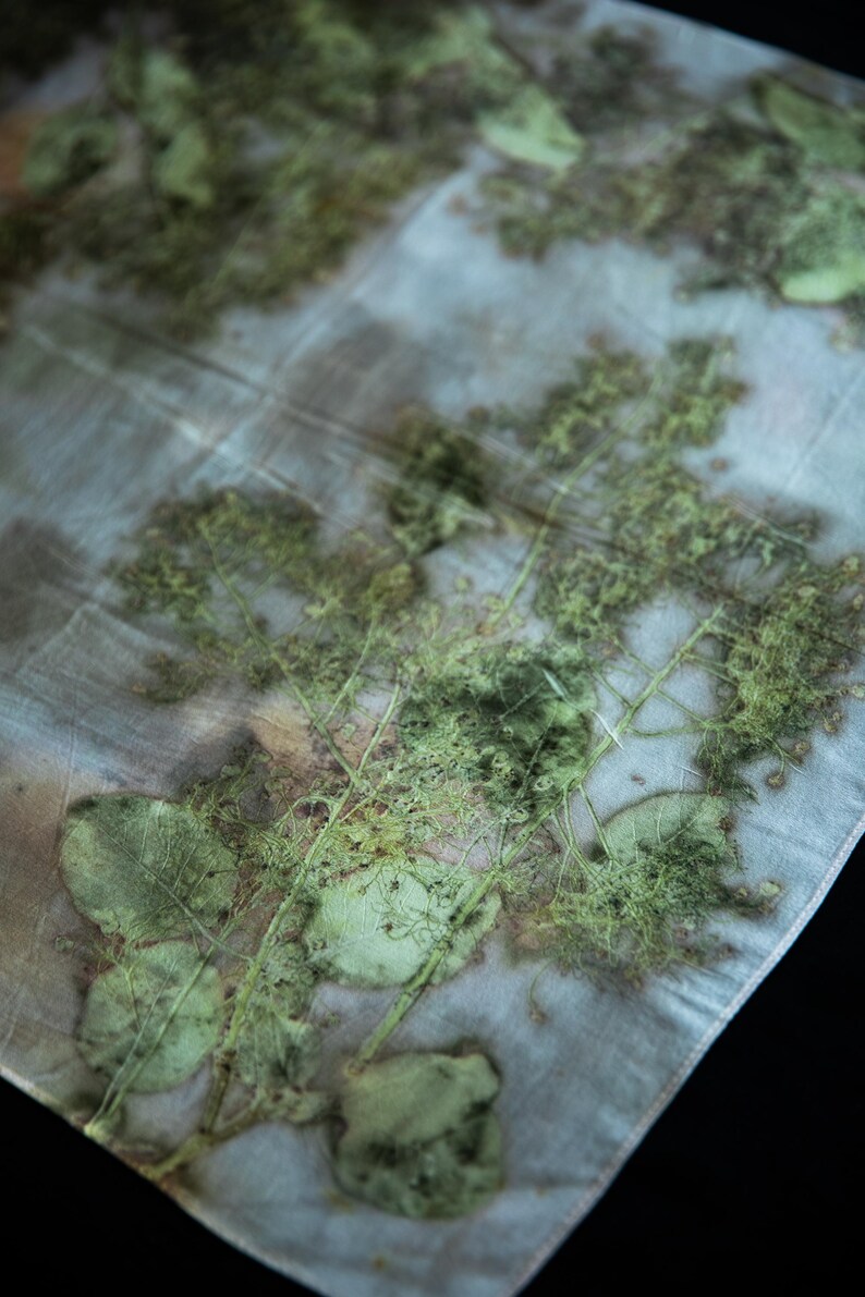 May include: A semi-sheer fabric featuring a botanical print with green leaf and fern silhouettes layered over a pale, muted blue and grey background. The textile displays organic, detailed plant imprints with visible veins and textures.
