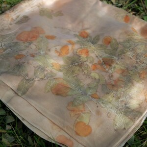 May include: A lightweight, semi-sheer scarf in a soft beige hue, featuring a botanical print of orange-toned fruit and green leaves. The fabric displays a natural, organic pattern created through plant-based dyeing, with visible leaf veins.