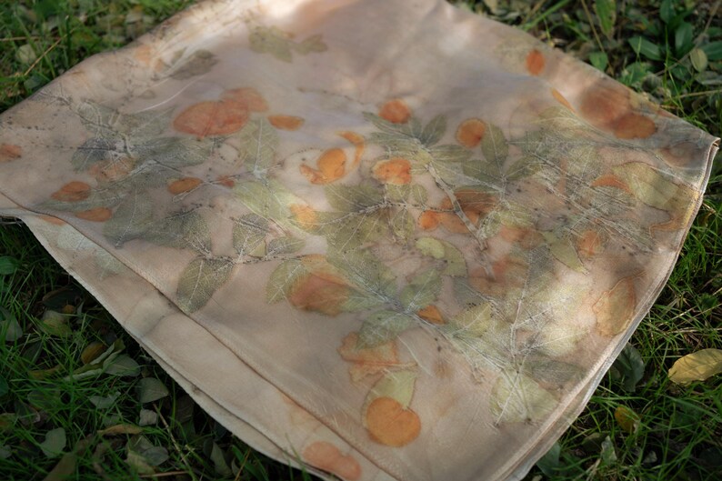 May include: A lightweight, semi-sheer scarf in a soft beige hue, featuring a botanical print of orange-toned fruit and green leaves. The fabric displays a natural, organic pattern created through plant-based dyeing, with visible leaf veins.