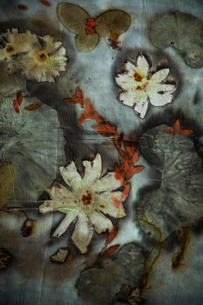 May include: A textile featuring a botanical print with pressed white flowers, large grey-green leaves, and a central orange-red branch. The fabric has a crinkled texture with dark, moody shadows and organic, earth-toned plant imprints throughout.