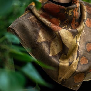 May include: A square silk scarf featuring a botanical print with dark brown, golden yellow, and burnt orange leaf patterns. The fabric displays a natural, organic aesthetic with detailed leaf silhouettes scattered across the surface in a repeating design.