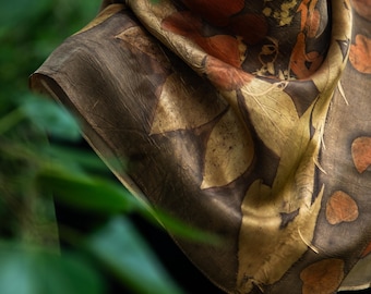 Puede incluir: Un pañuelo de seda cuadrado con un estampado botánico de hojas en tonos marrón oscuro, amarillo dorado y naranja quemado. La tela muestra una estética natural y orgánica con siluetas de hojas detalladas distribuidas en un patrón repetitivo.