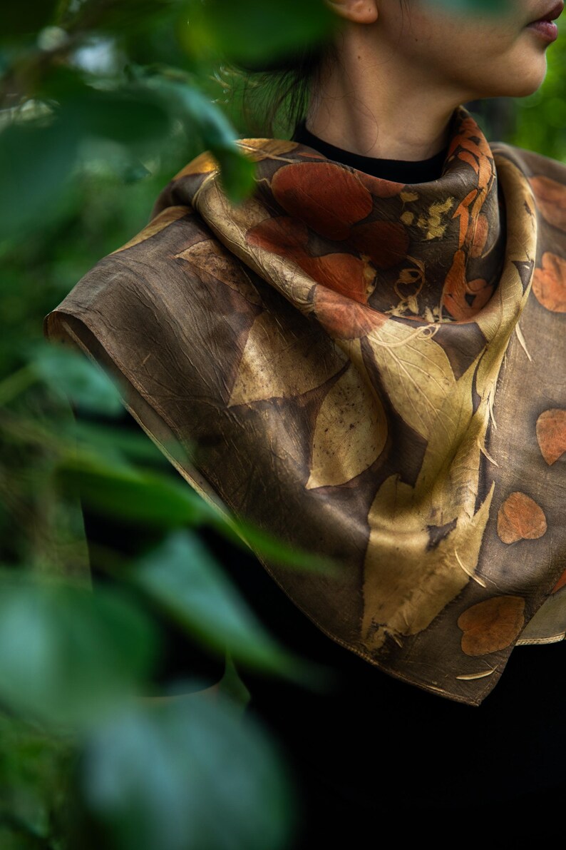 May include: A square silk scarf featuring a botanical print with dark brown, golden yellow, and burnt orange leaf patterns. The fabric displays a natural, organic aesthetic with detailed leaf silhouettes scattered across the surface in a repeating design.