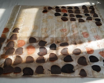 Puede incluir: Una pieza textil cuadrada que presenta una disposición en cuadrícula de impresiones circulares en forma de hoja de color marrón oscuro y naranja apagado sobre un fondo crema. La tela muestra una textura sutil y semitransparente con patrones botánicos.