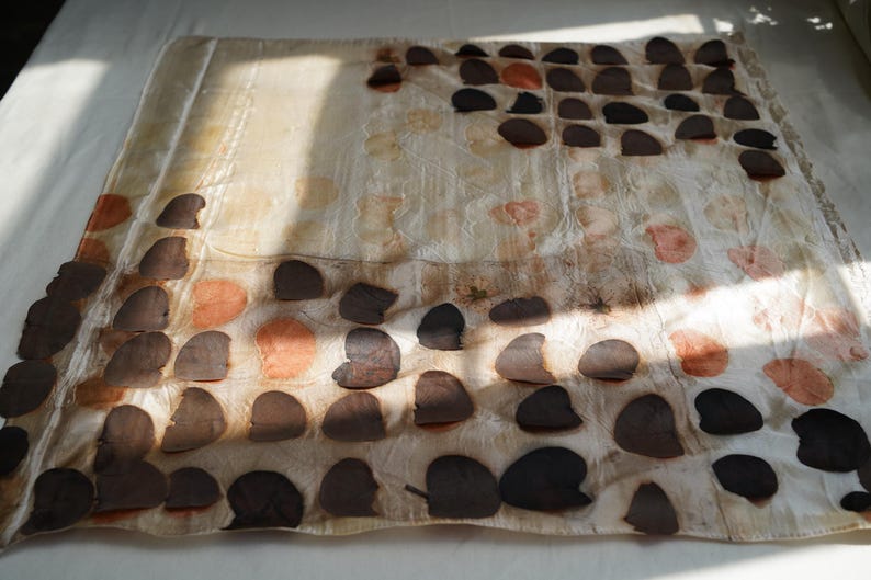 May include: A square textile piece featuring a grid-like arrangement of dark brown and muted orange circular leaf-like imprints on a cream-colored background. The fabric displays a subtle, semi-sheer texture with organic botanical patterns throughout.