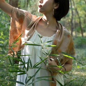 May include: A white sleeveless top with a gathered neckline paired with a beige silk scarf featuring botanical leaf prints in shades of orange and brown. The scarf is draped over the shoulders, showcasing organic patterns and a smooth fabric texture.