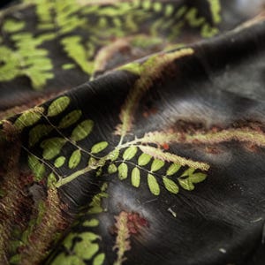 May include: A dark charcoal fabric featuring a botanical print with vibrant green fern-like leaves and reddish-brown textured accents. The material has a subtle, crinkled texture and drapes in soft folds, highlighting the detailed plant patterns.