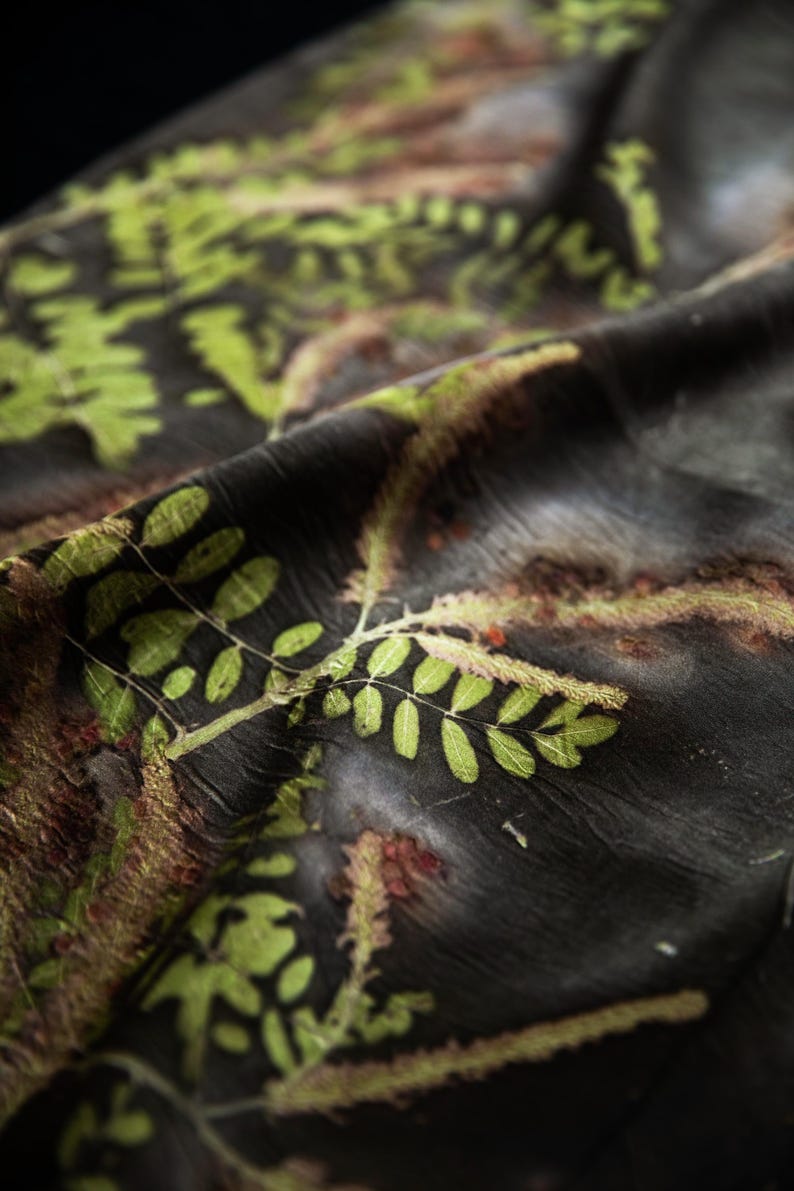 May include: A dark charcoal fabric featuring a botanical print with vibrant green fern-like leaves and reddish-brown textured accents. The material has a subtle, crinkled texture and drapes in soft folds, highlighting the detailed plant patterns.