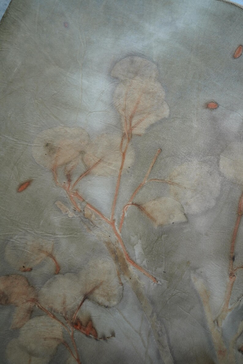 May include: A piece of fabric featuring a botanical print with muted sage green and gray tones. The design displays delicate, light-colored leaf and branch imprints with thin, reddish-brown stems and small, scattered orange-brown petal accents.