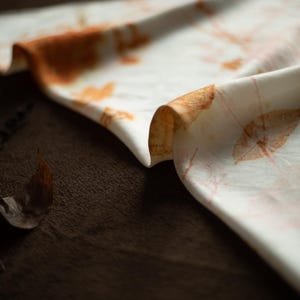 May include: A white fabric scarf featuring a botanical print with orange and brown leaf and stem patterns. The material has a smooth texture and is draped in soft folds, displaying a repeating natural design across the surface of the textile.