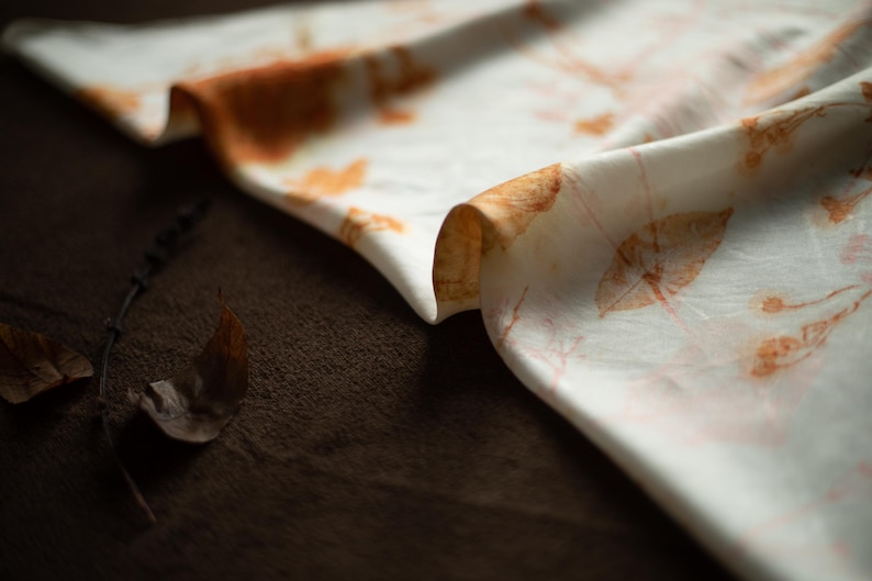May include: A white fabric scarf featuring a botanical print with orange and brown leaf and stem patterns. The material has a smooth texture and is draped in soft folds, displaying a repeating natural design across the surface of the textile.