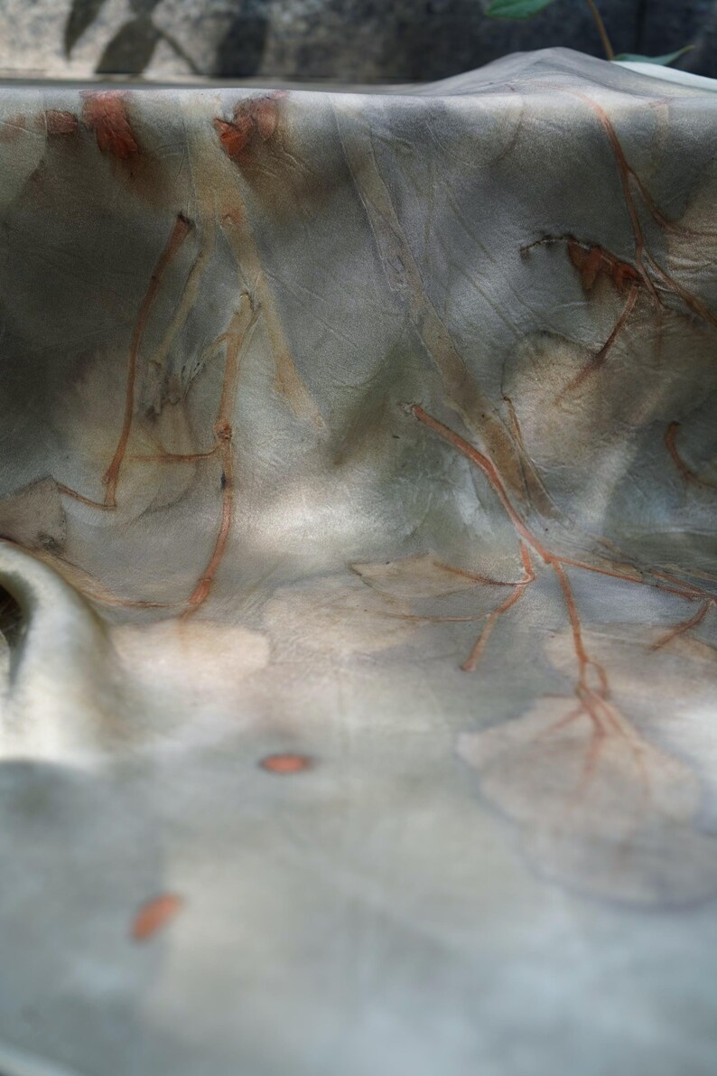 May include: A piece of fabric featuring a botanical print with muted gray and green tones. The surface displays intricate, rust-colored branch and leaf patterns that appear embedded into the material, creating a soft, organic, and textured visual effect.