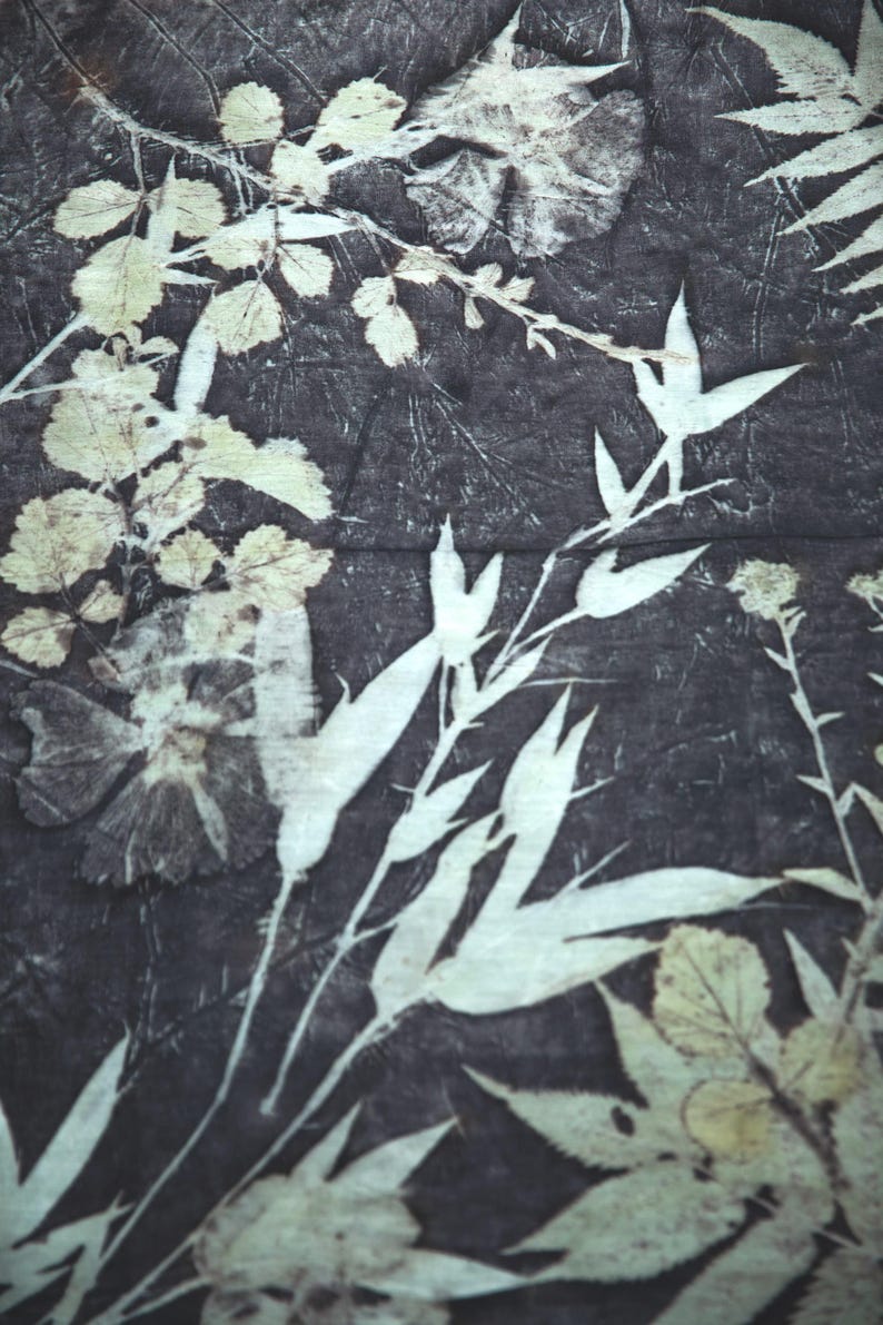 May include: A textile fabric featuring a dark charcoal gray background with a botanical print. The pattern consists of light cream-colored silhouettes of various leaves, stems, and flowers arranged in an overlapping, natural composition.