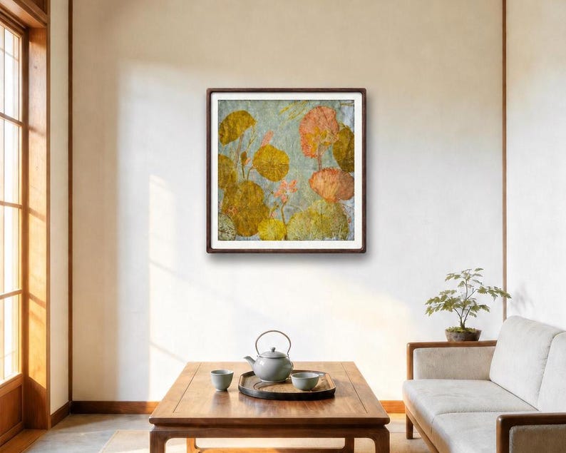 May include: A square framed wall art piece featuring a textured botanical illustration of golden-yellow and soft peach-colored lotus leaves and flowers against a light gray background, displayed in a dark brown wooden frame on a plain wall.