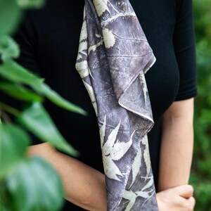 May include: A long, rectangular scarf featuring a dark grey and off-white botanical print with visible leaf and branch silhouettes. The fabric has a smooth texture and is draped vertically, showcasing a repeating pattern of foliage across the surface.