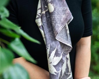 May include: A long, rectangular scarf featuring a dark grey and off-white botanical print with visible leaf and branch silhouettes. The fabric has a smooth texture and is draped vertically, showcasing a repeating pattern of foliage across the surface.