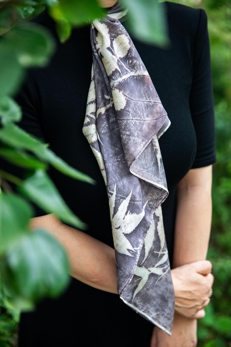 May include: A long, rectangular scarf featuring a dark grey and off-white botanical print with visible leaf and branch silhouettes. The fabric has a smooth texture and is draped vertically, showcasing a repeating pattern of foliage across the surface.
