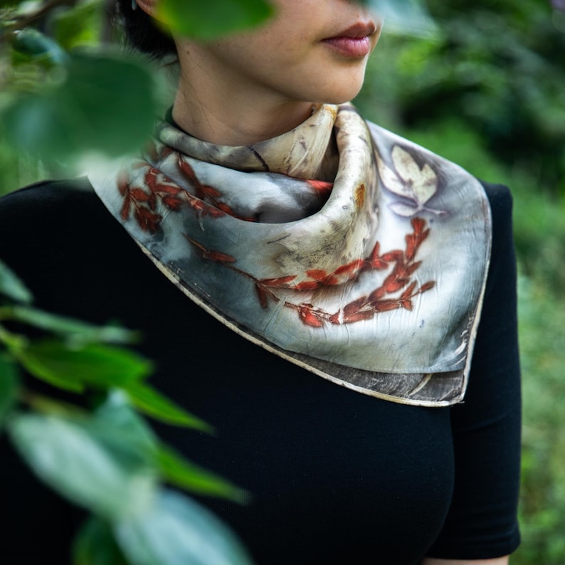 May include: A square silk scarf featuring a botanical print with muted grey and cream tones, accented by reddish-brown leaf and branch patterns. The scarf is folded and tied around the neck, displaying a smooth texture and natural plant-based designs.