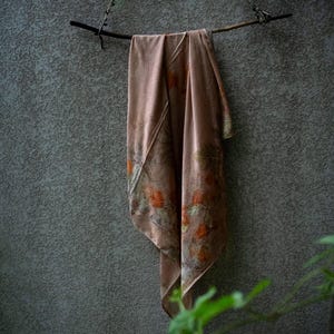 May include: A dusty rose-coloured scarf featuring a botanical print with orange and green leaf patterns. The scarf is draped over a dark, thin wooden branch that is suspended by a thin cord, creating a soft, flowing display of the fabric.