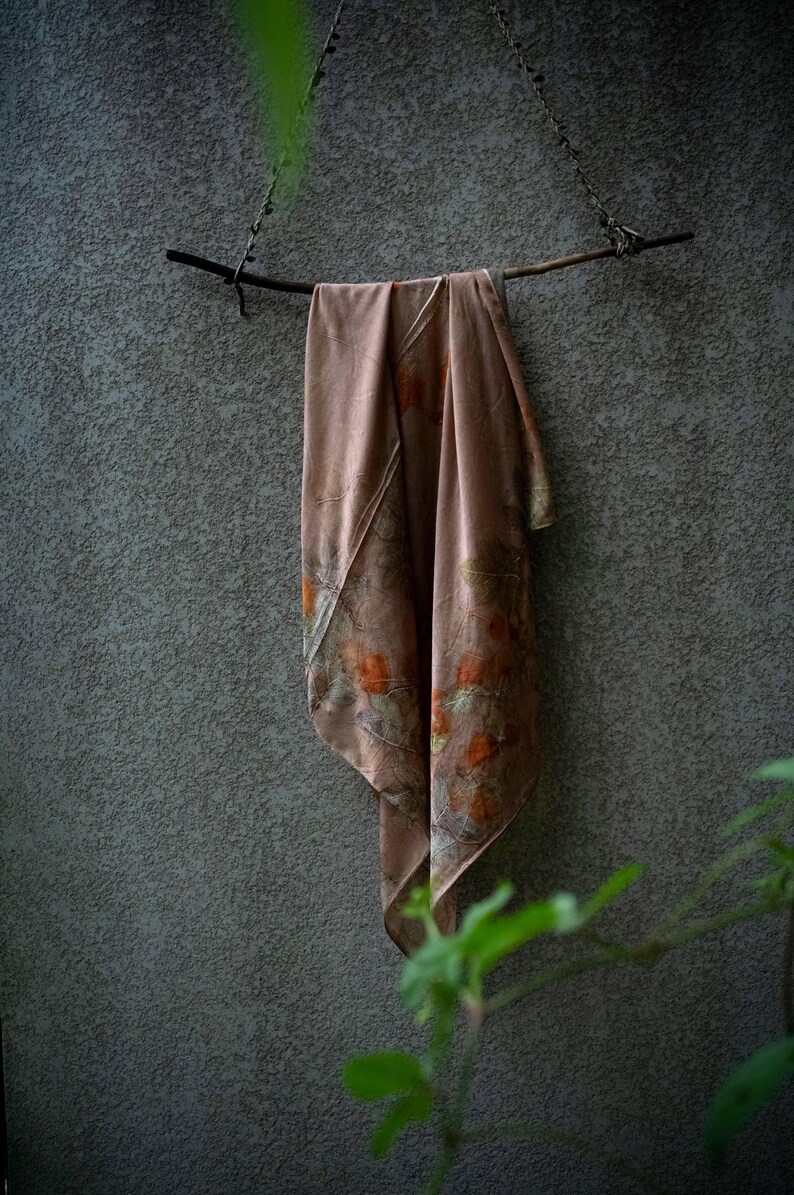 May include: A dusty rose-coloured scarf featuring a botanical print with orange and green leaf patterns. The scarf is draped over a dark, thin wooden branch that is suspended by a thin cord, creating a soft, flowing display of the fabric.