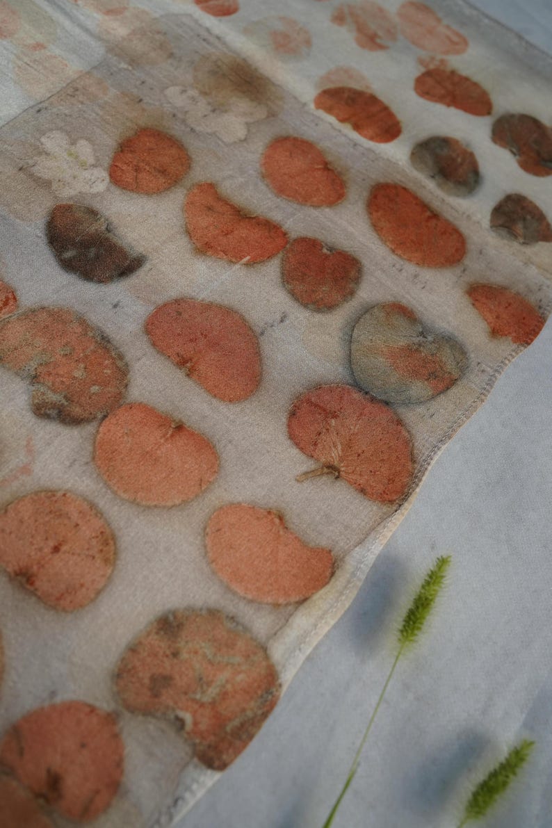 K&ouml;nnte beinhalten: Ein halbtransparenter, cremewei&szlig;er Stoffschal mit einem sich wiederholenden Muster aus botanischen Blattabdr&uuml;cken in Terrakotta-, Rost- und ged&auml;mpften Graut&ouml;nen. Das Textil weist eine nat&uuml;rliche Struktur mit sichtbaren Blattadern auf.