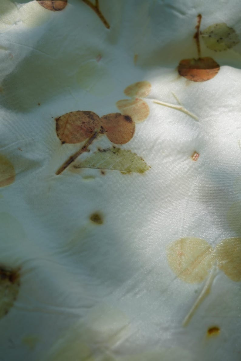 May include: A white fabric featuring a botanical print with scattered brown and pale green leaf imprints. The material has a smooth, slightly reflective surface with visible folds and creases, showcasing organic shapes of various foliage patterns.