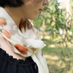 以下が含まれることがあります： オレンジと茶色の模様が入ったクリーム色のスカーフが首に巻かれています。スカーフは黒いトップスと組み合わせています。背景はぼやけており、緑の葉が見えます。