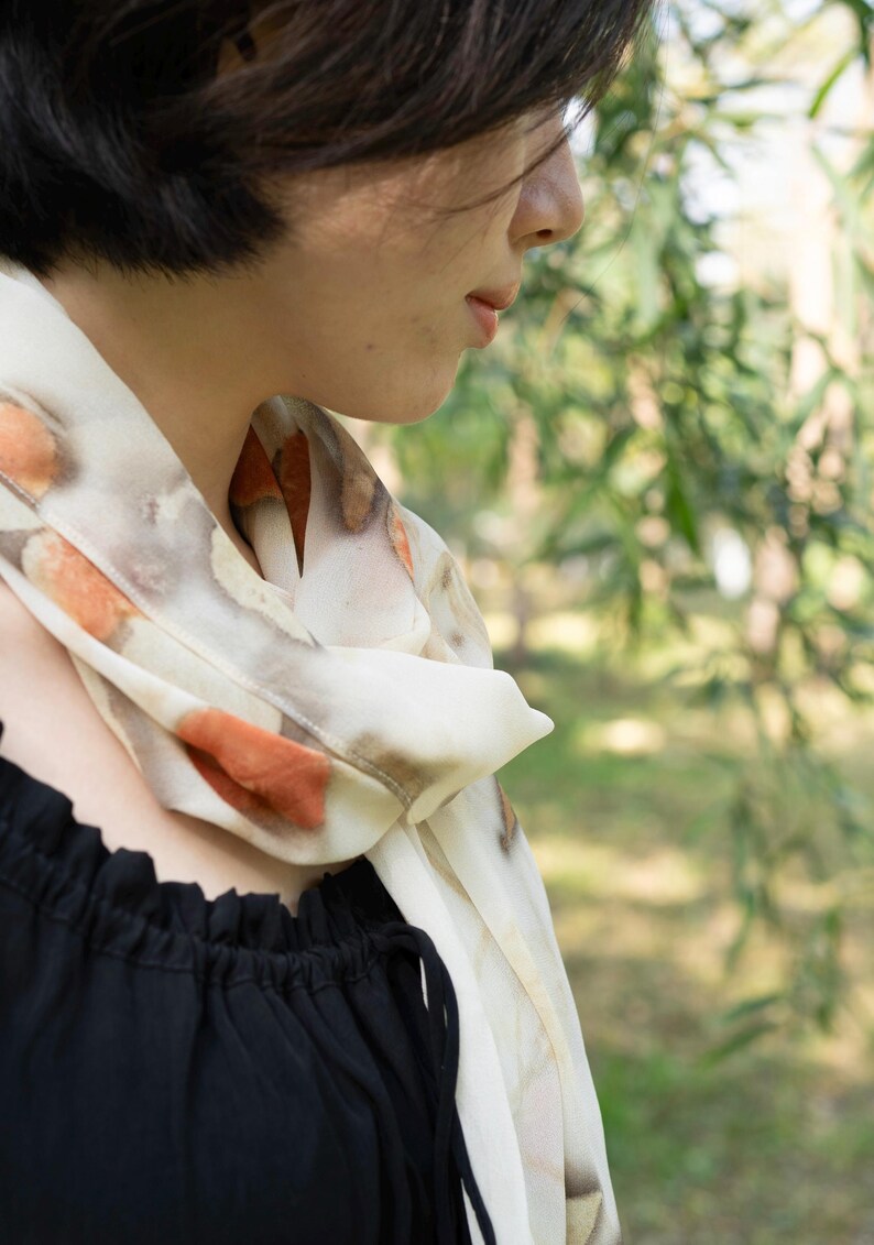 以下が含まれることがあります： オレンジと茶色の模様が入ったクリーム色のスカーフが首に巻かれています。スカーフは黒いトップスと組み合わせています。背景はぼやけており、緑の葉が見えます。