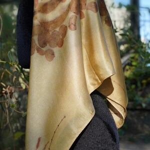 Pu&ograve; includere: Un foulard quadrato in seta di una tenue tonalit&agrave; giallo oro, caratterizzato da impronte botaniche di foglie e steli marroni. Il tessuto ha una finitura liscia e lucente con un drappeggio naturale, che mostra dettagliati motivi vegetali.