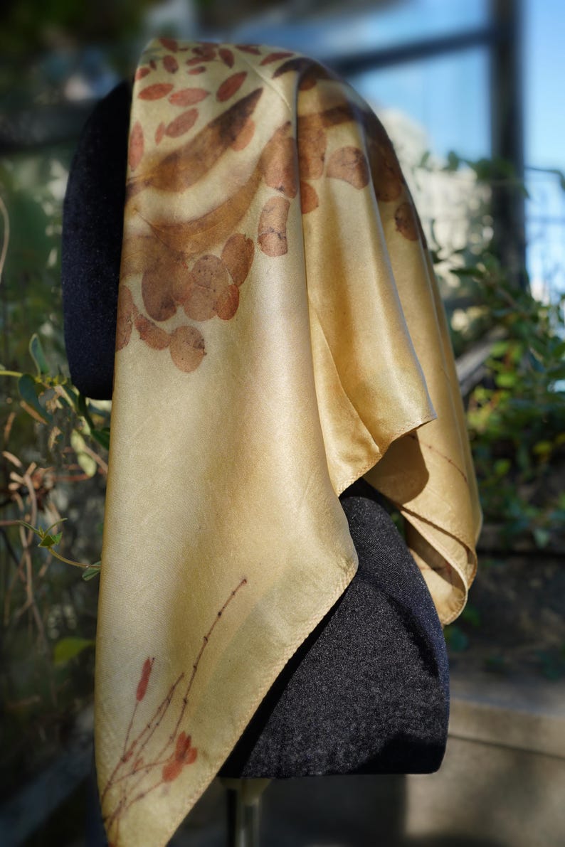 Pu&ograve; includere: Un foulard quadrato in seta di una tenue tonalit&agrave; giallo oro, caratterizzato da impronte botaniche di foglie e steli marroni. Il tessuto ha una finitura liscia e lucente con un drappeggio naturale, che mostra dettagliati motivi vegetali.