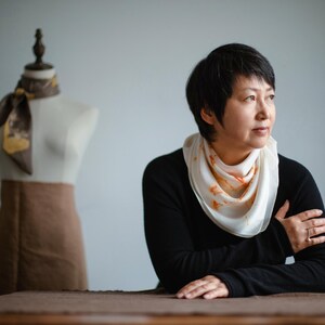 May include: A white silk scarf with orange botanical prints worn around the neck, paired with a black long-sleeved shirt. In the background, a mannequin displays a brown skirt and a dark brown silk scarf with a gold-toned botanical pattern.