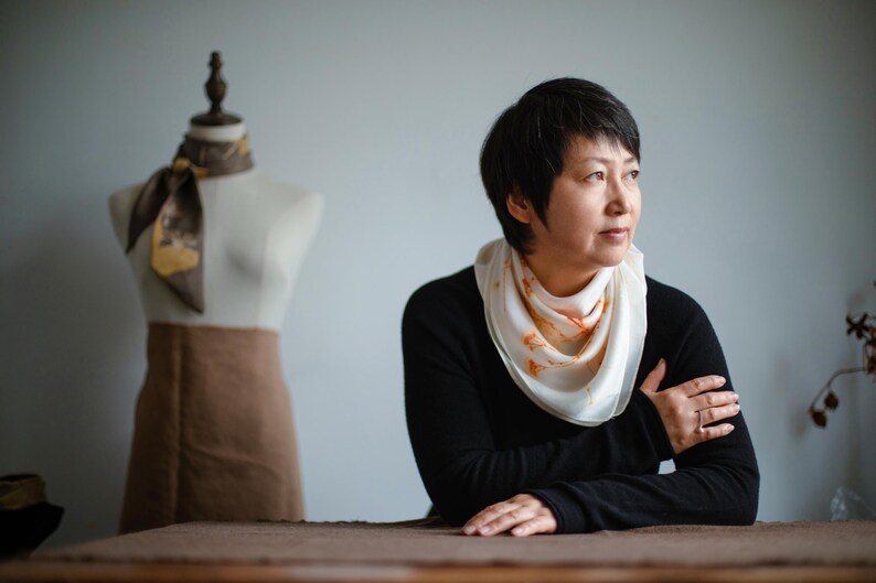 May include: A white silk scarf with orange botanical prints worn around the neck, paired with a black long-sleeved shirt. In the background, a mannequin displays a brown skirt and a dark brown silk scarf with a gold-toned botanical pattern.